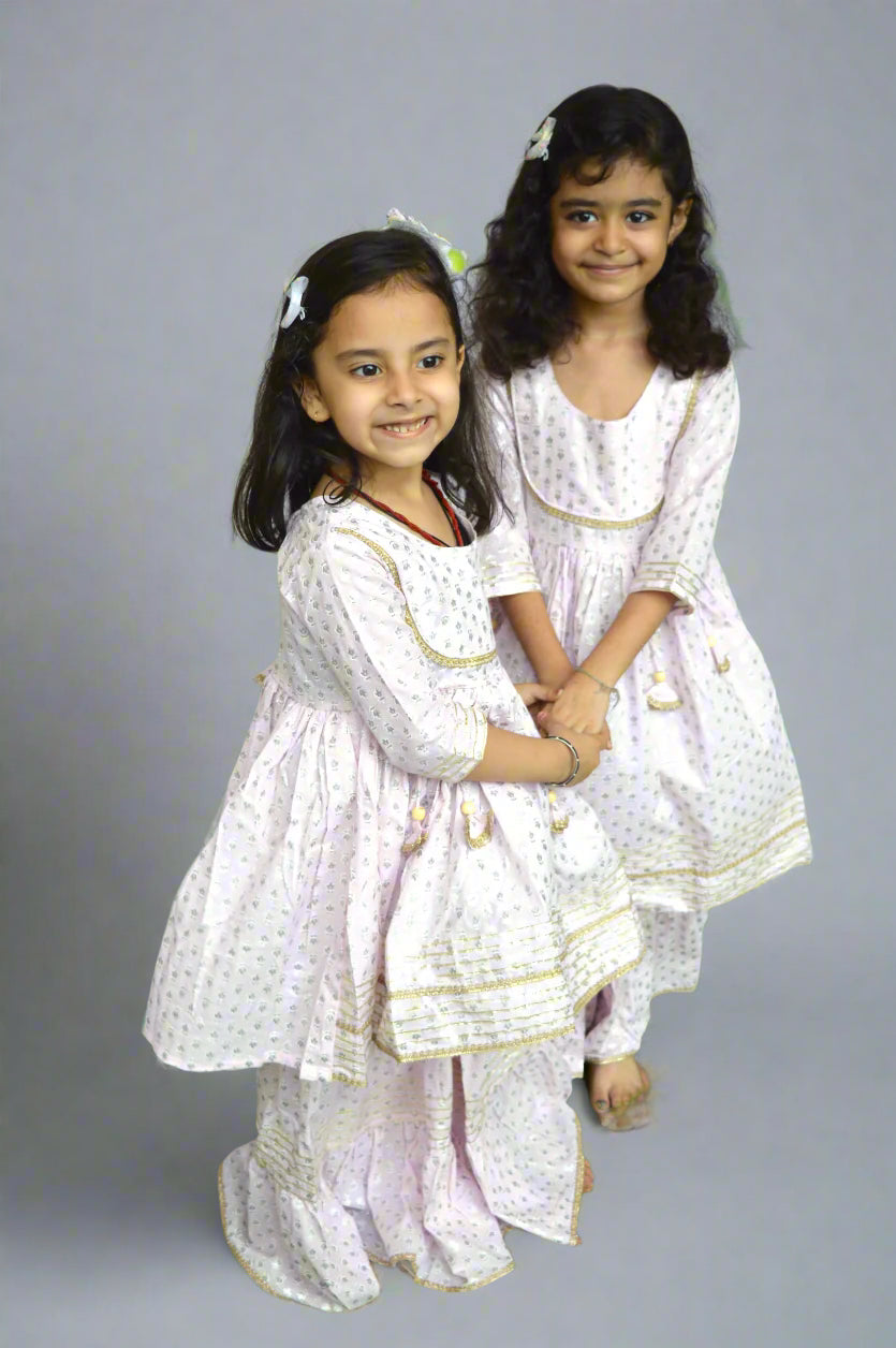 Two young girls in matching white dresses standing on a grassy surface with a white curtain background.